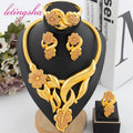 Gold jewelry set including necklace, earrings, and bracelet on a black stand with flowers and books in the background.