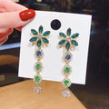 Pair of floral earrings with green and blue stones on a white card held by a hand with red nail polish.