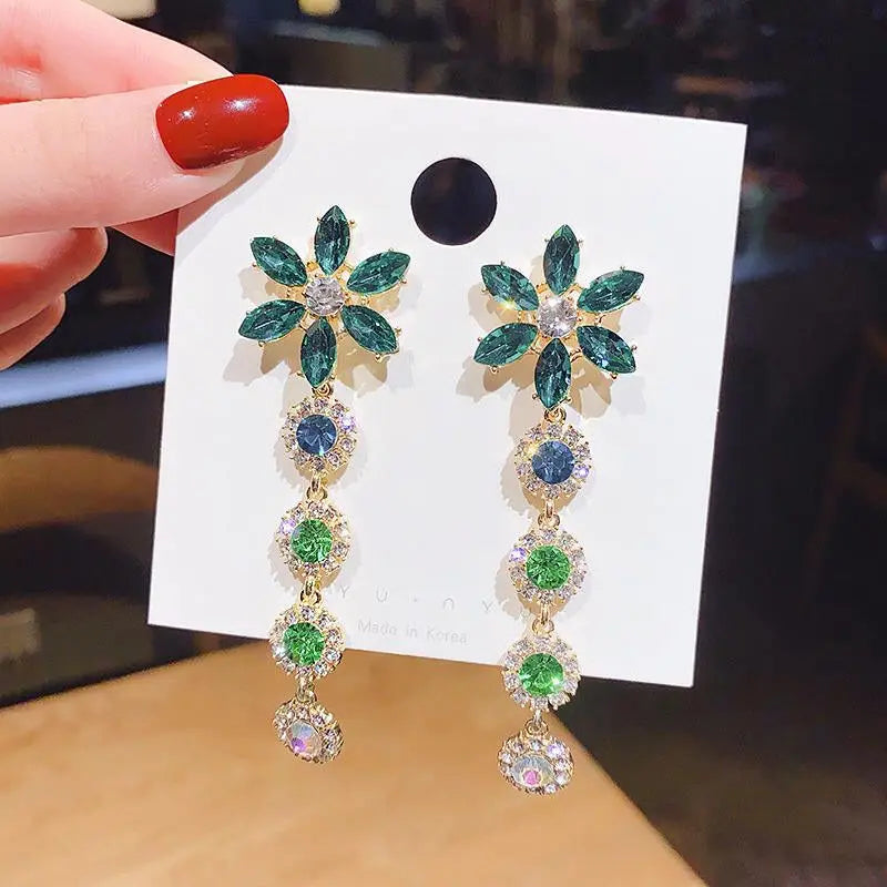 Pair of floral earrings with green and blue stones on a white card held by a hand with red nail polish.