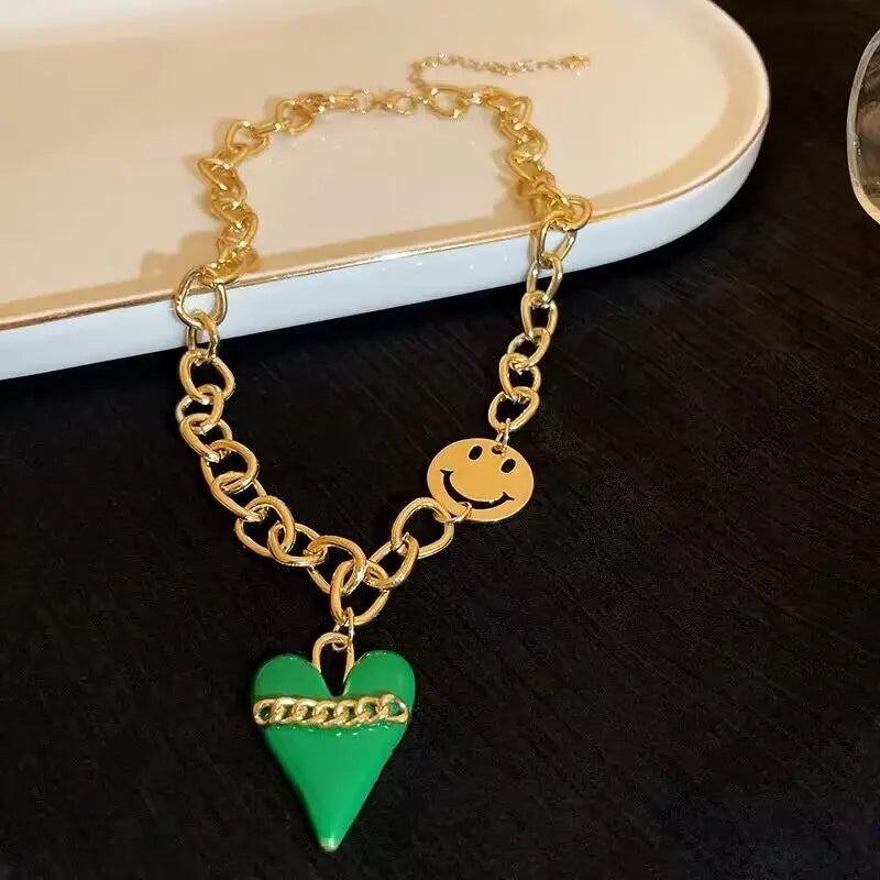 Gold chain bracelet with a green heart and smiley face charm on a dark surface.