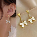 Gold bow-shaped earrings worn by a person, with close-up on ear and side view on beige fabric.